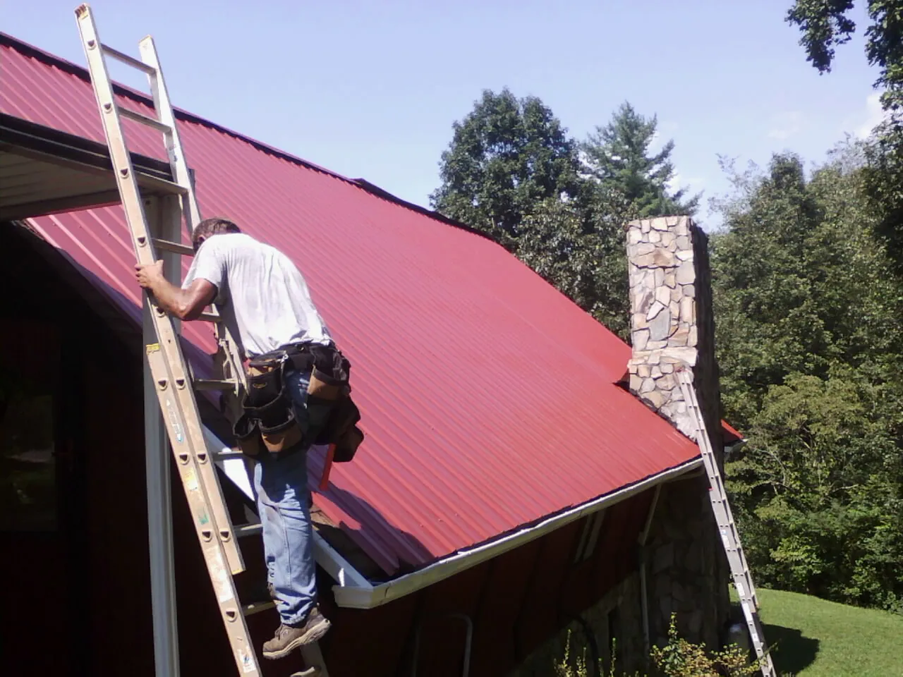 Roofer on a ladder beside a red metal roof and stone chimney — Soffit & Fascia Repair by Roofing Brothers in New Preston Marble Dale